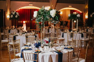 Traditional romantic wedding reception decor, hanging draped lights, gold chiavari chairs, tall floral centerpiece with white hydrangeas, red and pink roses, greenery, navy blue linens | Tampa Bay wedding planner Special Moments Events | Wedding Venue St. Pete Coliseum