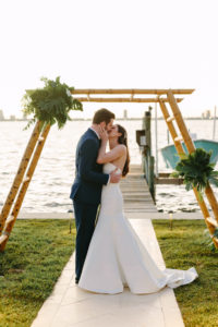 Modern Tropical Bride and Groom Kissing Under Bamboo Ladder Arch with Palm Leaves Arrangements, Waterfront Wedding Photo | Tampa Bay Wedding Planner, Florist and Designer John Campbell Weddings