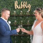 Bride and Groom Cheering with Champagne Flutes in front of Greenery Bubbly Champagne Wall | Tampa Bay Wedding Planner Coastal Coordinating | Wedding Rentals Outside the Box | Wedding Hair and Makeup Femme Akoi Beauty Studio | Wedding Dress Truly Forever Bridal
