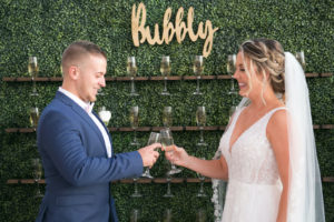 Bride and Groom Cheering with Champagne Flutes in front of Greenery Bubbly Champagne Wall | Tampa Bay Wedding Planner Coastal Coordinating | Wedding Rentals Outside the Box | Wedding Hair and Makeup Femme Akoi Beauty Studio | Wedding Dress Truly Forever Bridal