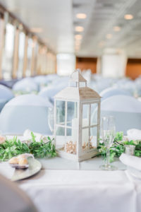 Indoor Wedding Reception at Tampa Venue Yacht Starship | White Reception Tables with Greenery Garland Runners and White Lantern Centerpieces | White Wedding Chair Covers with Silver Grey Sash Bow | Silver Charger Plate with White Rolled Napkin by Kate Ryan Event Rentals