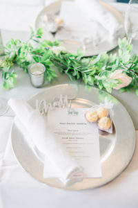 White Reception Tables with Greenery Garland Runners and Silver Charger Plate with White Rolled Napkin, Menu Card, and Chocolate Candy Favors