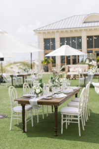 Garden-style beachside wedding reception decor, wooden feasting farm table with white linen table runner, white chairs, floral centerpieces | Tampa Bay wedding planner Parties A'la Carte | Clearwater Beach wedding venue Sandpearl Resort | Kate Ryan Event Rentals