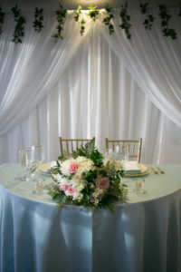 Elegant Modern Wedding Reception Decor, Sweetheart Table with Dusty Blue Linens, Gold Chiavari Chairs, Lush White and Pink Floral with Greenery Arrangement, White Background Draping and Hanging Greenery Leaves | Tampa Bay Wedding Photographer Carrie Wildes | Wedding Planner Coastal Coordinating | Wedding Florist Brides N Blooms