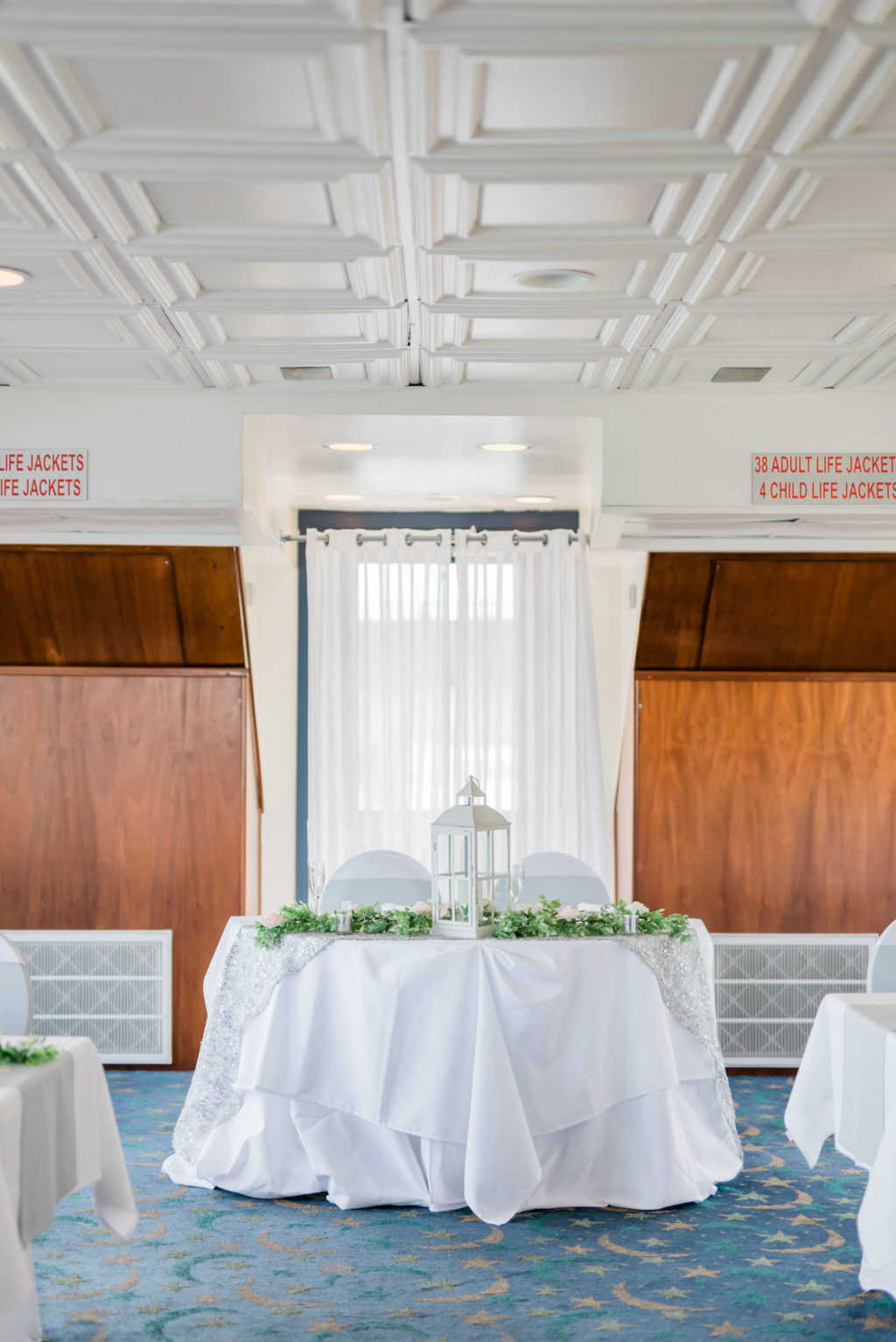 Indoor Wedding Reception at Tampa Venue Yacht Starship | White Sweetheart Table with Greenery Garland Runners and White Lantern Centerpieces | White Wedding Chair Covers with Silver Grey Sash Bow by Kate Ryan Event Rentals