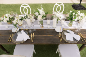 Garden-style beachside wedding reception decor, wooden farm feasting table, clear and gold rimmed chargers, white linens and table runner, gold silverware, white roses and greenery floral centerpieces, candles in hurricane vases, white chairs | Tampa Bay Wedding Planner Parties A'la Carte | Kate Ryan Event Rentals