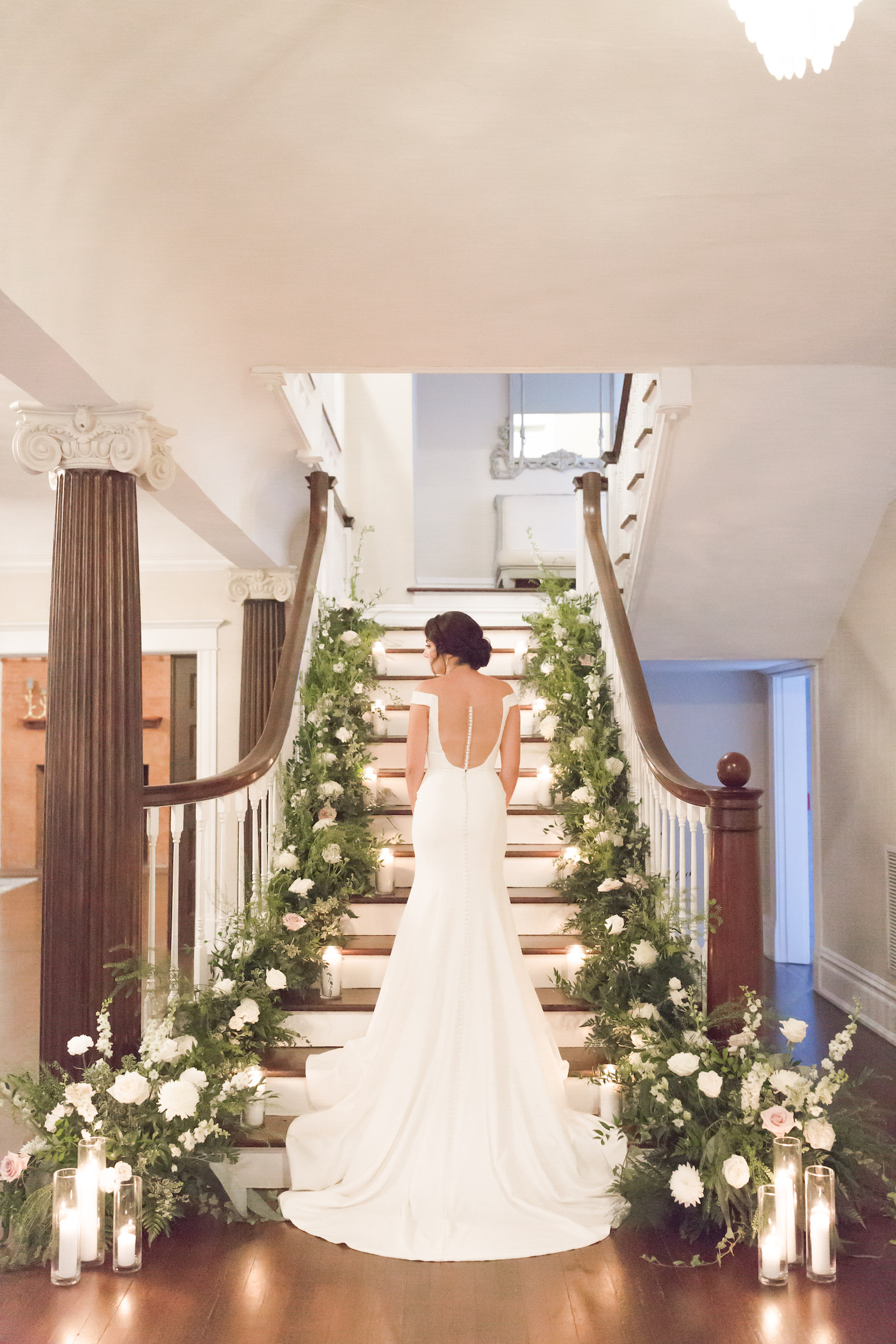 Classic bride wearing open back and cap sleeve wedding dress on staircase of historic Tampa wedding venue The Orlo, lush greenery and white floral arrangements lining staircase | Tampa Bay wedding planner Elegant Affairs by Design