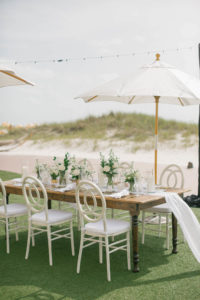 Garden-style beachside wedding reception decor, wooden farm feasting table with white table runner, white chairs, umbrellas, and floral centerpieces | Tampa Bay wedding planner Parties A'la Carte | Kate Ryan Event Rentals | Clearwater Beach Wedding Venue Sandpearl Resort