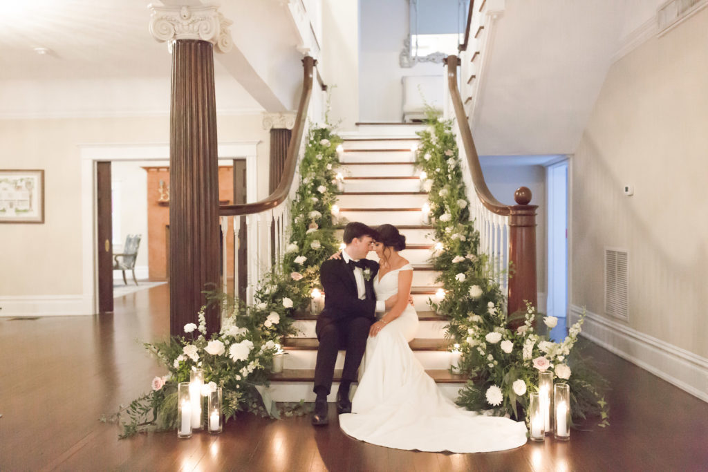 Classic bride and groom sitting on staircase of historic wedding venue The Orlo, Lush greenery and white floral arrangements lining staircase | Tampa Bay wedding planner and design Elegant Affairs by Design