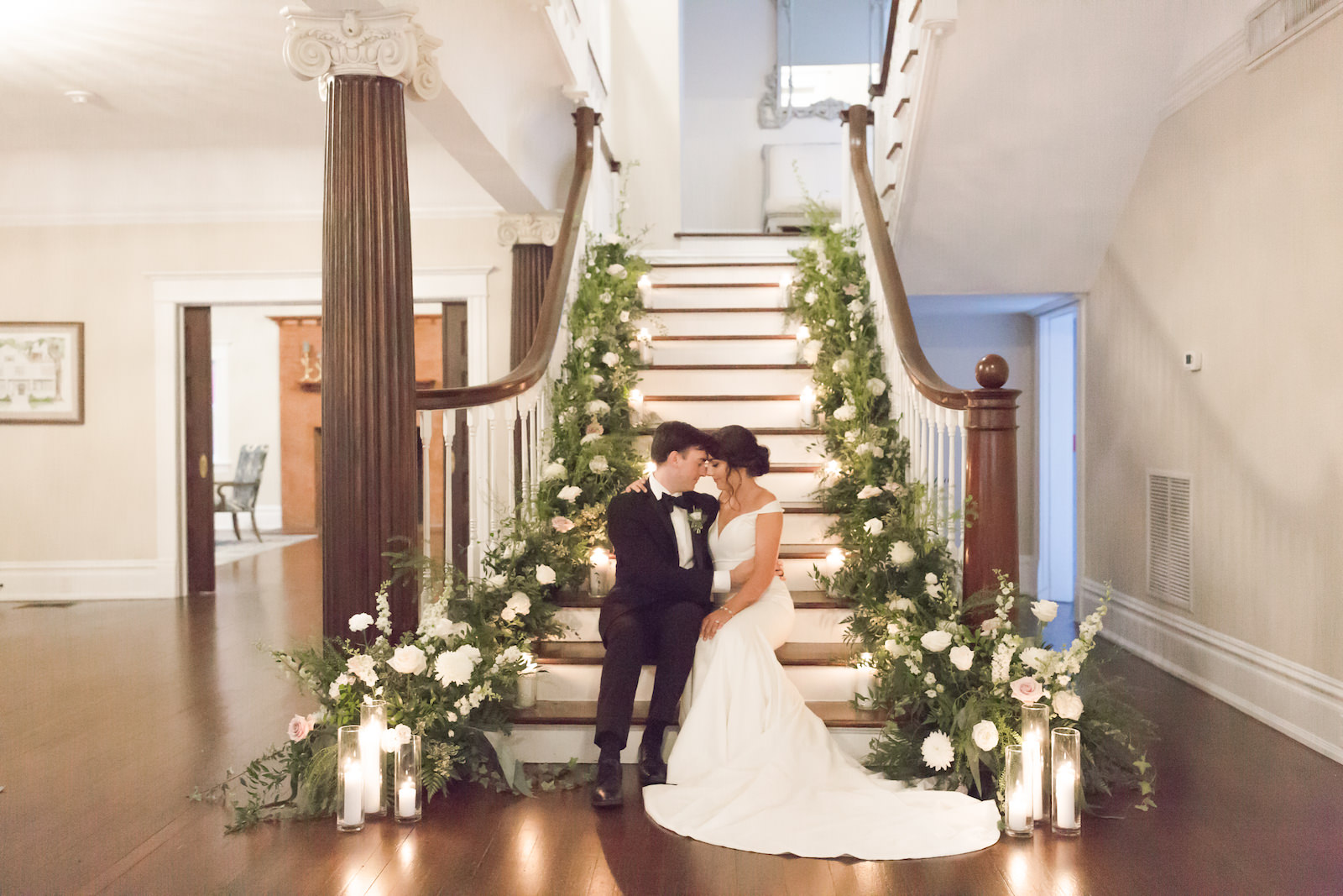 Classic bride and groom sitting on staircase of historic wedding venue The Orlo, Lush greenery and white floral arrangements lining staircase | Tampa Bay wedding planner and design Elegant Affairs by Design