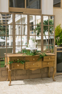 Garden-style wedding reception decor, farmhouse wooden dresser and mirror with three tier white wedding cake garnished with greenery | Tampa Bay Wedding Planner Parties A'la Carte