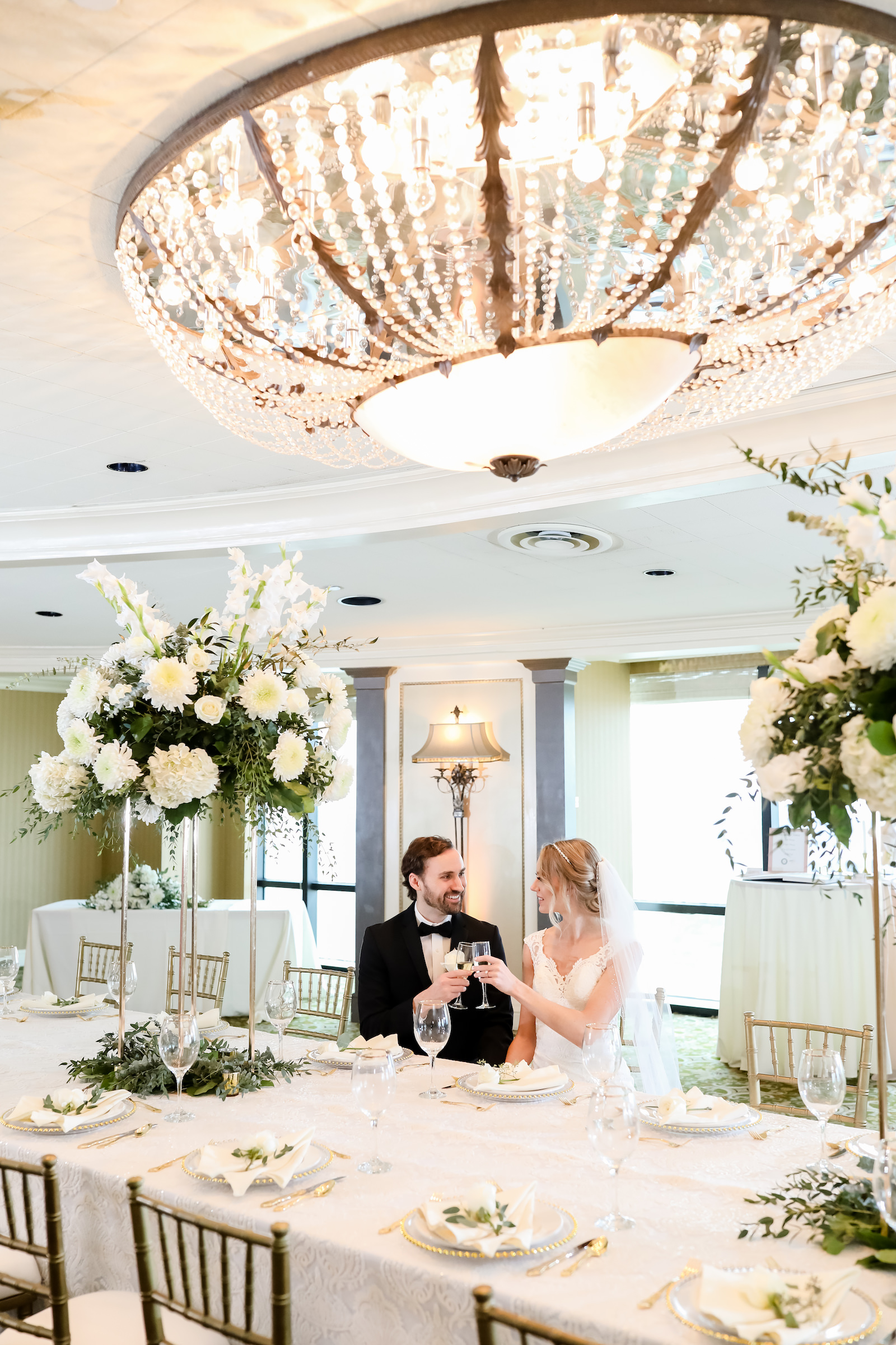 Classic Bride and Groom Cheering a Glass of Champagne During Wedding Reception, Long Feasting Table with White Linens, Tall Floral Centerpieces with Lush White and Greenery Floral Centerpieces | Tampa Bay Wedding Planner Elegant Affairs by Design | Wedding Venue The Tampa Club | Wedding Rentals Outside the Box Event Rentals | Wedding Photography Lifelong Photography Studio | Styled Shoot