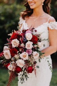 Bride Wearing Romantic Lace Cap Sleeve Maggie Sottero Wedding Dress Holding Berry Colored, Red, Blush and Purple Floral Arrangement