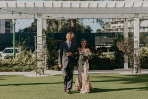 Bride Walking Down Aisle with Father during Outdoor Clearwater Wedding Ceremony | Boho Gold Bronze Embroidered Overlay Wedding Dress Bridal Gown with Illusion Sleeves and Train | Tropical Boho Bridal Bouquet