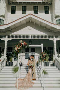 Outdoor Staircase Wedding Ceremony Bride and Groom Portrait on Belleview Inn Staircase | Boho Gold Bronze Embroidered Overlay Wedding Dress Bridal Gown with Illusion Sleeves and Train | Groom in Grey Khaki Linen Suit | Tropical Boho Bridal Bouquet and White Arch Backdrop with Palm Frond Leaf Coral Orange Pink and Greenery Floral Arrangements | Amber McWhorter Photography