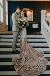 Indoor Bride and Groom Portrait on Staircase | Boho Gold Bronze Embroidered Overlay Wedding Dress Bridal Gown with Illusion Sleeves and Train | Groom in Grey Khaki Linen Suit | Tropical Boho Bridal Bouquet | Amber McWhorter Photography