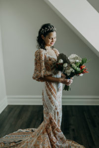 Indoor Bridal Portrait by Window with Natural Light | Boho Gold Bronze Embroidered Overlay Wedding Dress Bridal Gown with Illusion Sleeves and Train | Tropical Boho Bridal Bouquet with White Roses Orange Orchids and Palm Frond Leaves