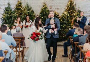 Tampa Christmas Wedding Ceremony with Lit Christmas Trees Backdrop | Long Sleeve Lace Plunging V Neck Tulle Ballgown Wedding Dress Bridal Gown with Long Cathedral Veil | Groom Wearing Navy Blue Jacket with Black Satin Lapel and Bow Tie | Red Burgundy and Gold Rose Bridal Bouquet