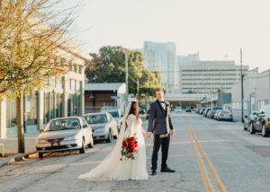 Outdoor Street Bride and Groom Portrait | Downtown Tampa Heights Christmas Wedding | Long Sleeve Lace Plunging V Neck Tulle Ballgown Wedding Dress Bridal Gown with Long Cathedral Veil | Groom Wearing Navy Blue Jacket with Black Satin Lapel and Bow Tie | Red Burgundy and Gold Rose Bridal Bouquet