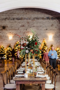 Indoor Tampa Christmas Wedding Reception with Wood Farm Feasting Tables and Tall Gold Centerpieces of Red White and Gold Cafe au Lait Roses and Winter Greenery and Berries | Lit Christmas Tree Backdrop