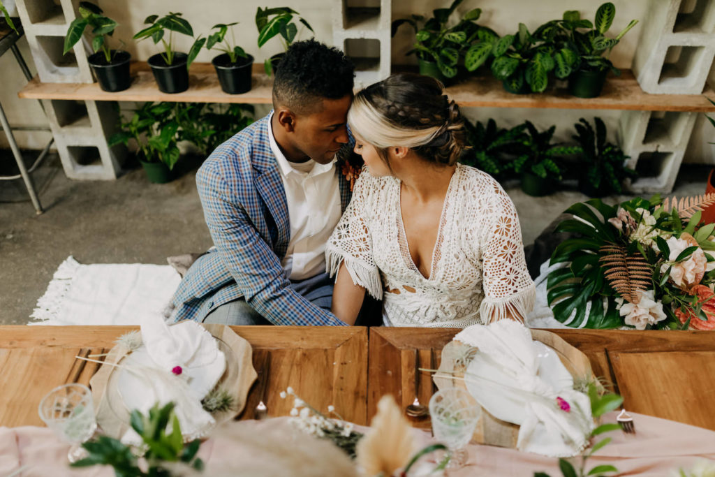 Boho Neutral Styled Shoot | Bride in Vintage Lace and Fringe Short Sleeve V Neckline Wedding Dress, Groom in Blue Plaid Suit Sitting at Wooden Table with Chargers, White Linens, Greenery Plants | Unique Wedding Planner Elope Tampa Bay | Amber McWhorter Photography | Wooden Chargers and Rose Gold Flatware Rentals Kate Ryan Event Rentals