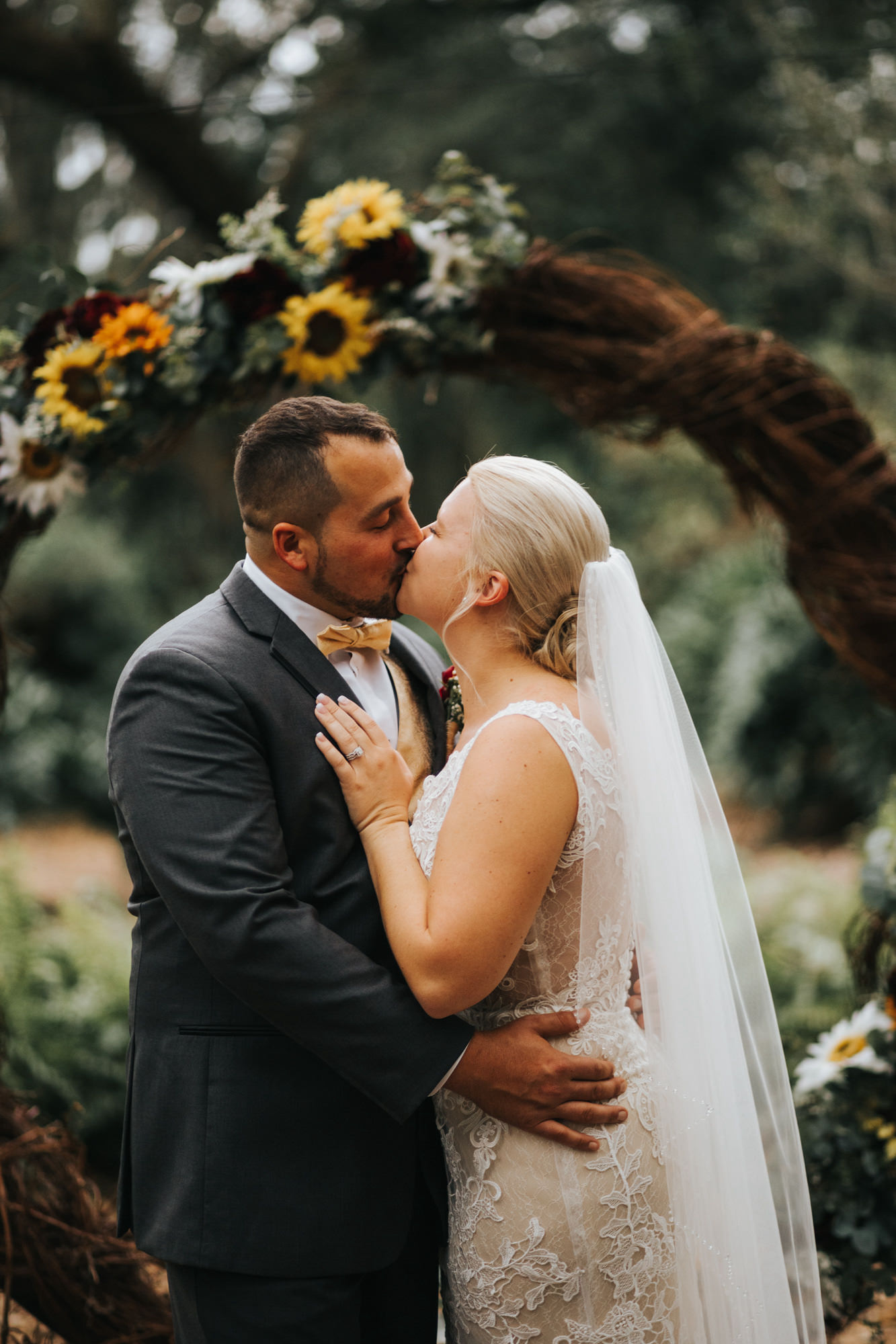 Bride and Groom First Kiss during Rustic Dover Burgundy and Yellow Sunflower Outdoor Wedding Ceremony with Round Grapevine Branch Arch | V Neck Champagne Lace Bridal Gown Wedding Dress from Sarasota Dress Shop Truly Forever Bridal | Groom in Classic Charcoal Grey Gray Suit Tux with Sunflower Boutonniere