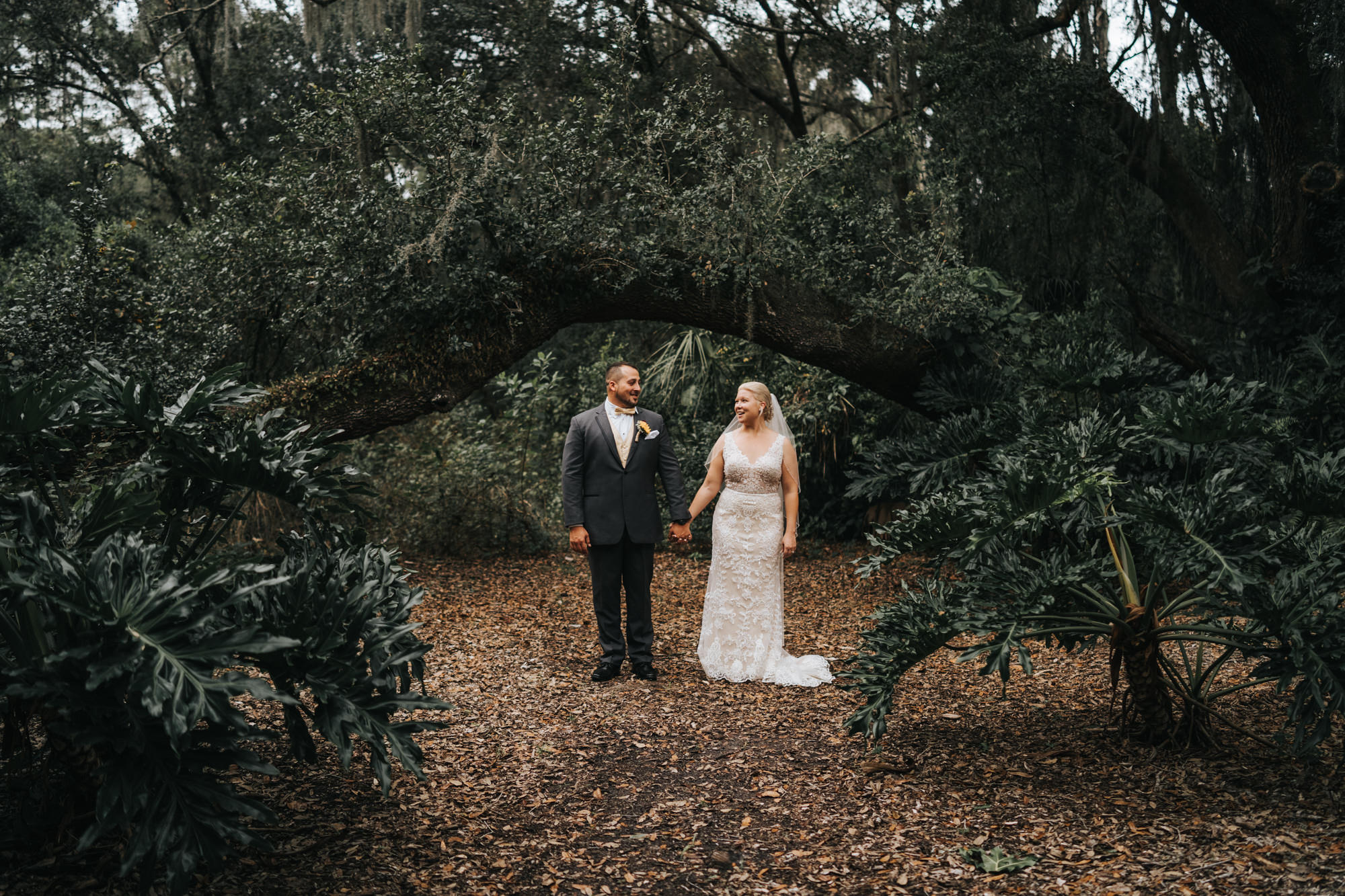 Bride and Groom Outdoor Portrait in Dover Florida with Oak Trees and Greenery | V Neck Champagne Lace Bridal Gown Wedding Dress from Sarasota Dress Shop Truly Forever Bridal | Groom in Classic Charcoal Grey Gray Suit Tux with Sunflower Boutonniere