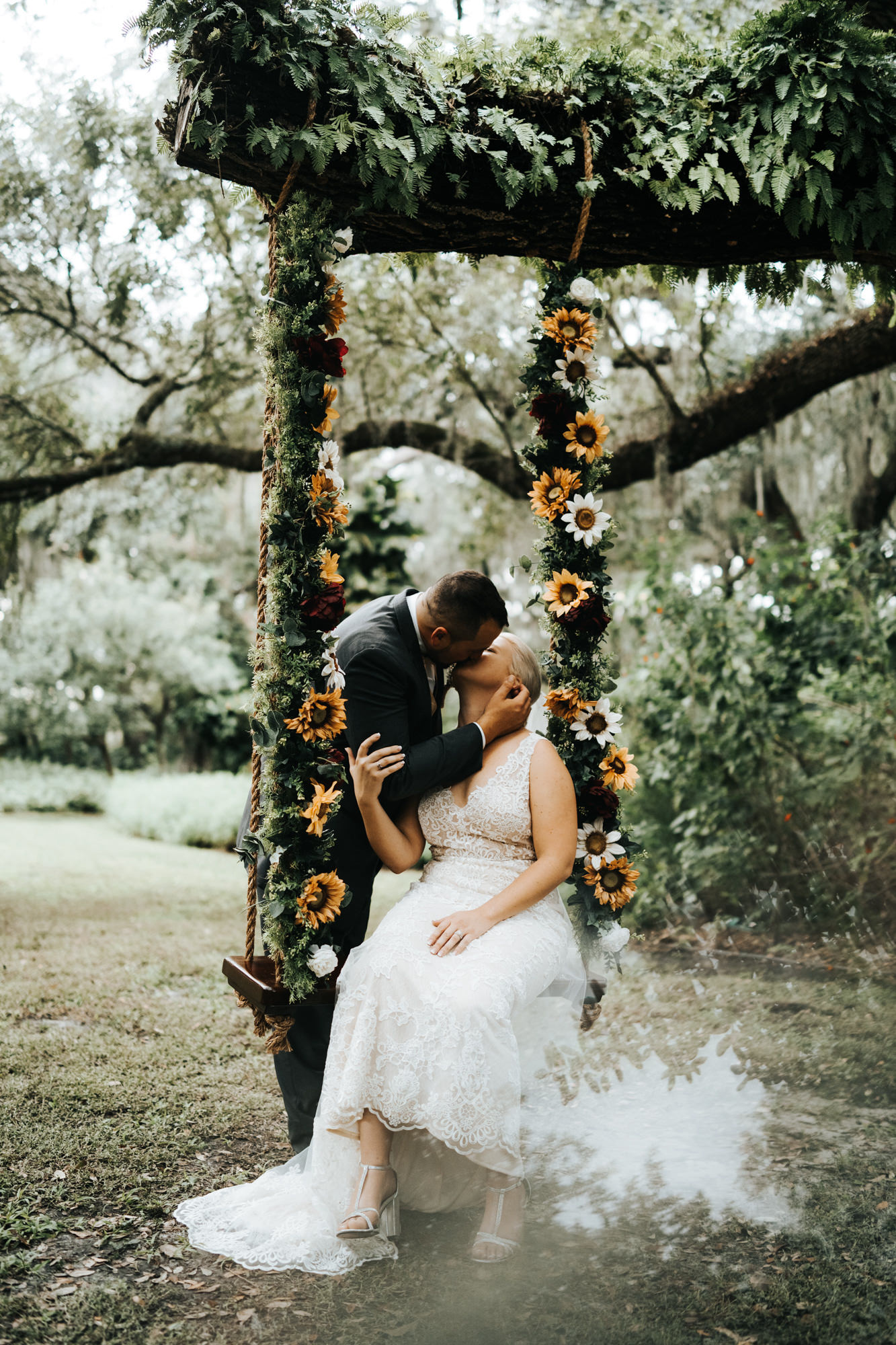 Bride and Groom Outdoor Portrait on Tree Swing with Greenery and Sunflowers | V Neck Champagne Lace Bridal Gown Wedding Dress from Sarasota Dress Shop Truly Forever Bridal | Groom in Classic Charcoal Grey Gray Suit Tux with Sunflower Boutonniere