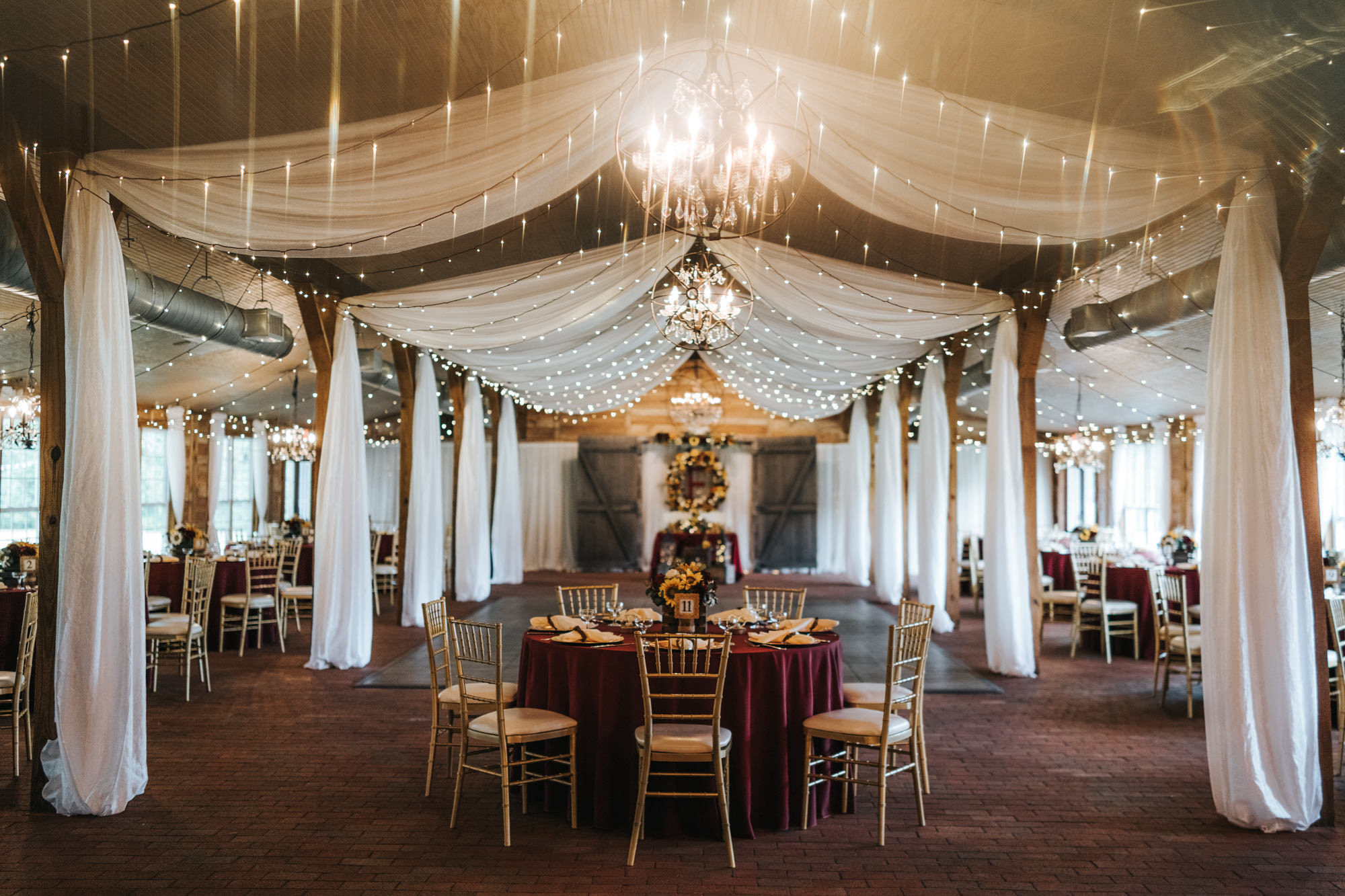 Rustic Dover Burgundy and Yellow Sunflower Wedding Barn Reception with String Lights and Ceiling Draping | Round Reception Tables with Burgundy Maroon Red Linen Table Cloths and Gold Chiavari Chairs