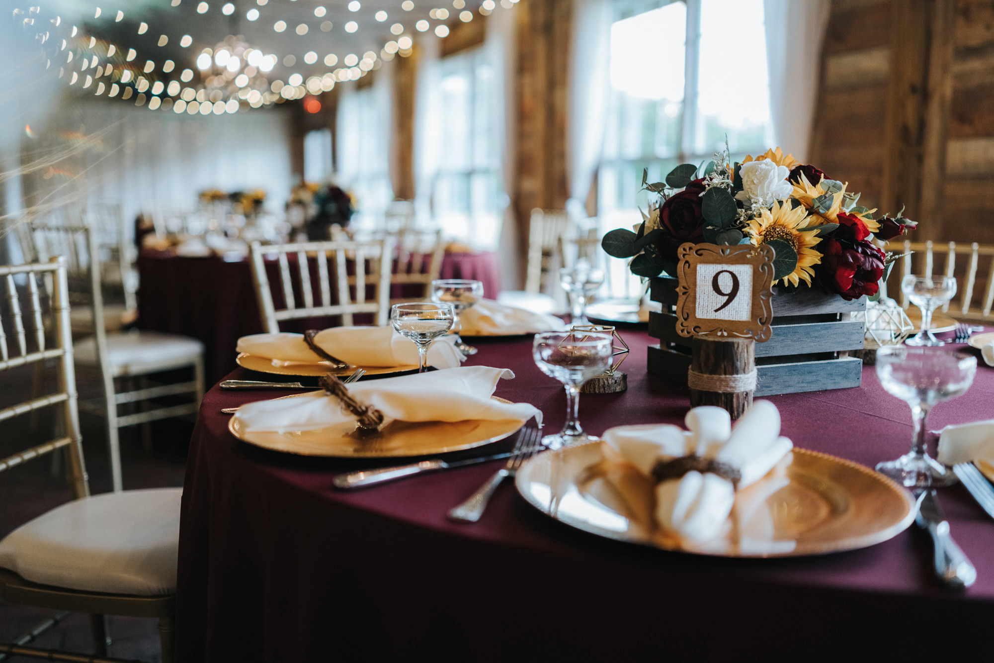 Rustic Dover Burgundy and Yellow Sunflower Wedding Barn Reception with String Lights and Ceiling Draping | Round Reception Tables with Burgundy Maroon Red Linen Table Cloths and Gold Chiavari Chairs | Wood Crate Centerpiece with Red Roses and Yellow Sunflowers with Gold Charger Plates and Wood Frame Table Numbers