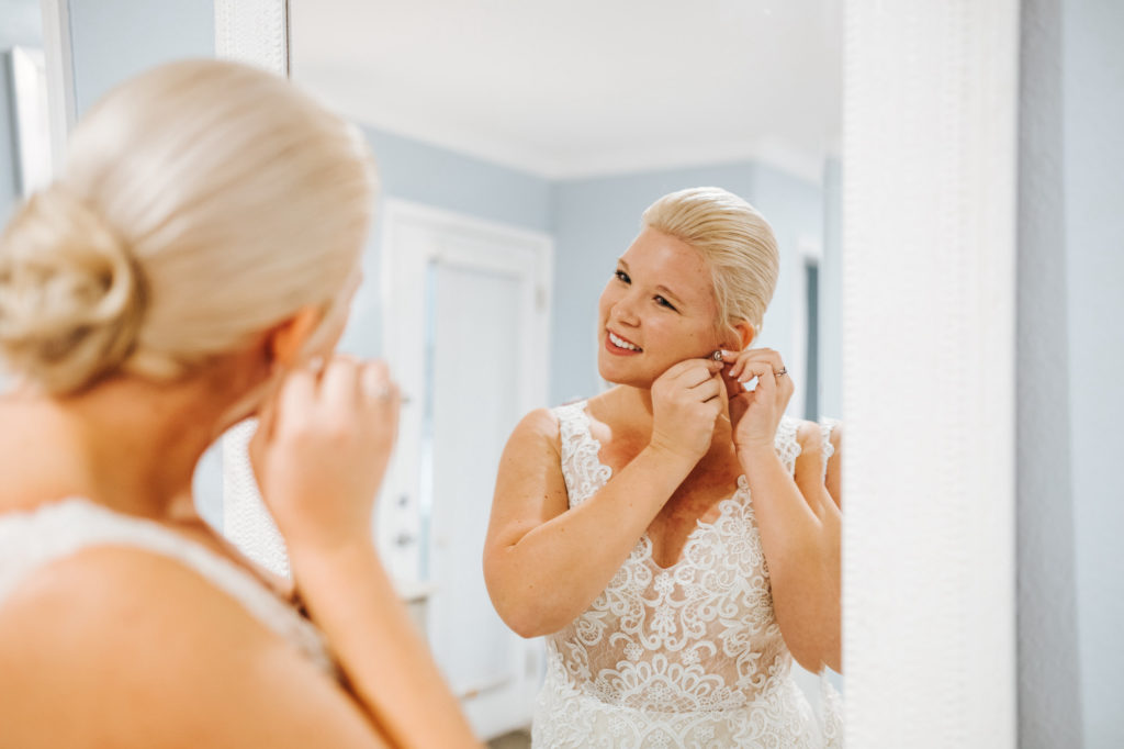 Bride Putting on Earrings Looking in Mirror | V Neck Champagne Lace Bridal Gown Wedding Dress from Sarasota Dress Shop Truly Forever Bridal | Low Bun Chignon Bridal Hairstyle