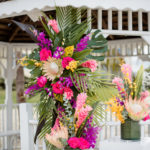 Tropical Florida Wedding Ceremony Decor, Alter Floral Arrangement, Bright Pink Exotic Flowers, Purple Stems, Blush King Proteas, with Green Palms and Monstera Leafs | Tampa Bay Vow Renewal and Microwedding Planner Perfecting The Plan | St. Petersburg Private Beachfront Venue Isla Del Sol