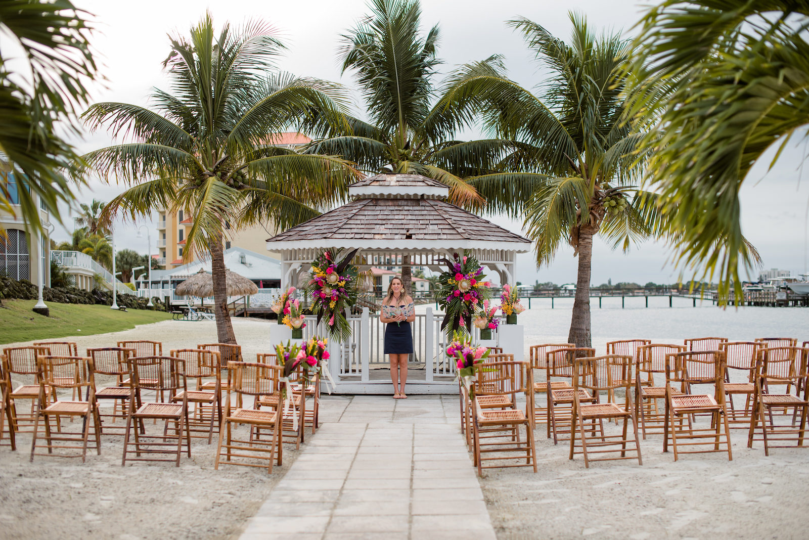 Tropical Florida Wedding Ceremony and Decor, Bamboo Folding Chairs, Lilly Pulitzer Themed Floral Arrangements, Gazebo Alter with Bright Pink Exotic Flowers, Purple Stems, Blush King Proteas, with Green Palms and Monstera Leafs | Tampa Bay Vow Renewal and Micro wedding Planner Perfecting The Plan | St. Petersburg Private Beachfront Venue Isla Del Sol | St. Pete Beach DJ Grant Hemond & Associates