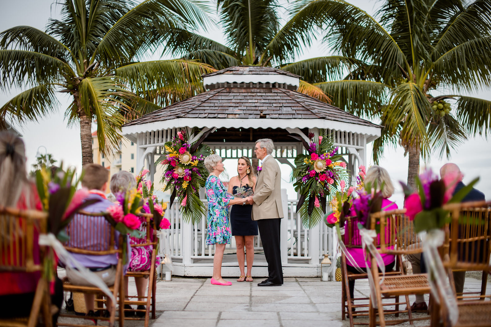 Florida Husband and Wife Hold Hands at Tropical Wedding Vow Renewal Ceremony with Decor, Bamboo Folding Chairs, Lilly Pulitzer Themed Floral Arrangements, Gazebo Alter with Bright Pink Exotic Flowers, Purple Stems, Blush King Proteas, with Green Palms and Monstera Leafs | Tampa Bay Vow Renewal and Micro wedding Planner Perfecting The Plan | St. Petersburg Private Beachfront Venue Isla Del Sol | St. Pete Beach DJ Grant Hemond & Associates