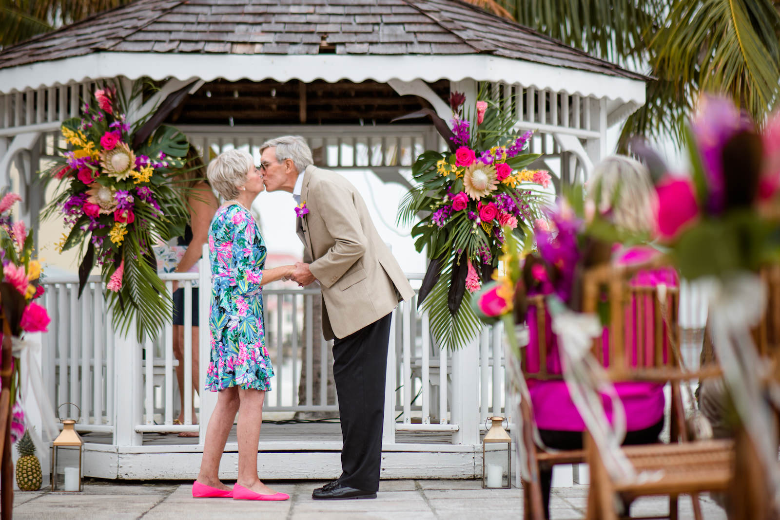 Florida Husband and Wife Kiss at Tropical Wedding Vow Renewal Ceremony with Decor, Bamboo Folding Chairs, Lilly Pulitzer Themed Floral Arrangements, Gazebo Alter with Bright Pink Exotic Flowers, Purple Stems, Blush King Proteas, with Green Palms and Monstera Leafs | Tampa Bay Vow Renewal and Micro wedding Planner Perfecting The Plan | St. Petersburg Private Beachfront Venue Isla Del Sol | St. Pete Beach DJ Grant Hemond & Associates