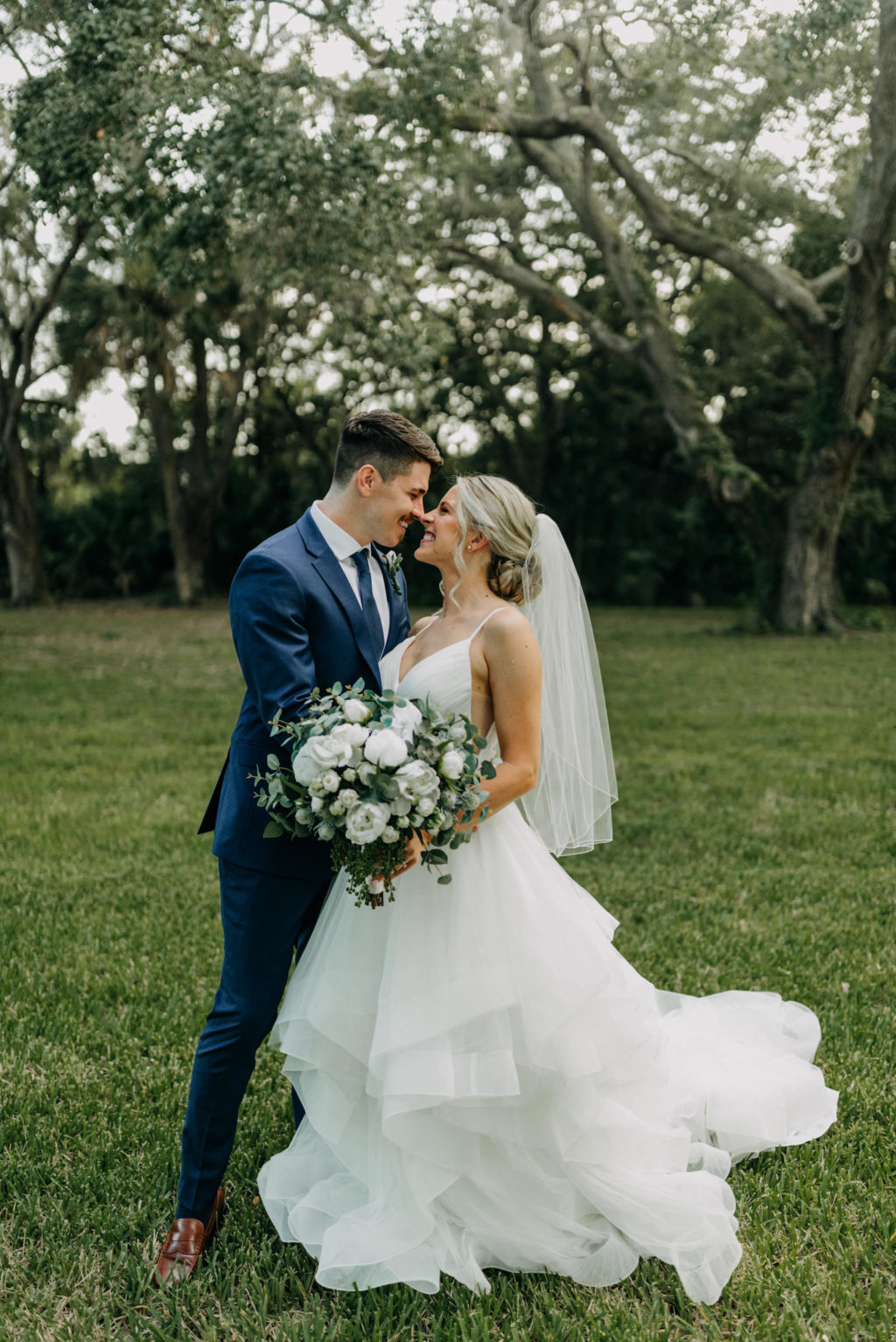 Classic Bride with Hair in Bun in Horsehair Ruffle Skirt V Neckline Wedding Dress Holding White and Greenery Eucalyptus Floral Bouquet Kissing Groom in Blue Suit | Tampa Bay Wedding Photographer Amber McWhorter Photography | Wedding Hair and Makeup Femme Akoi Beauty Studio