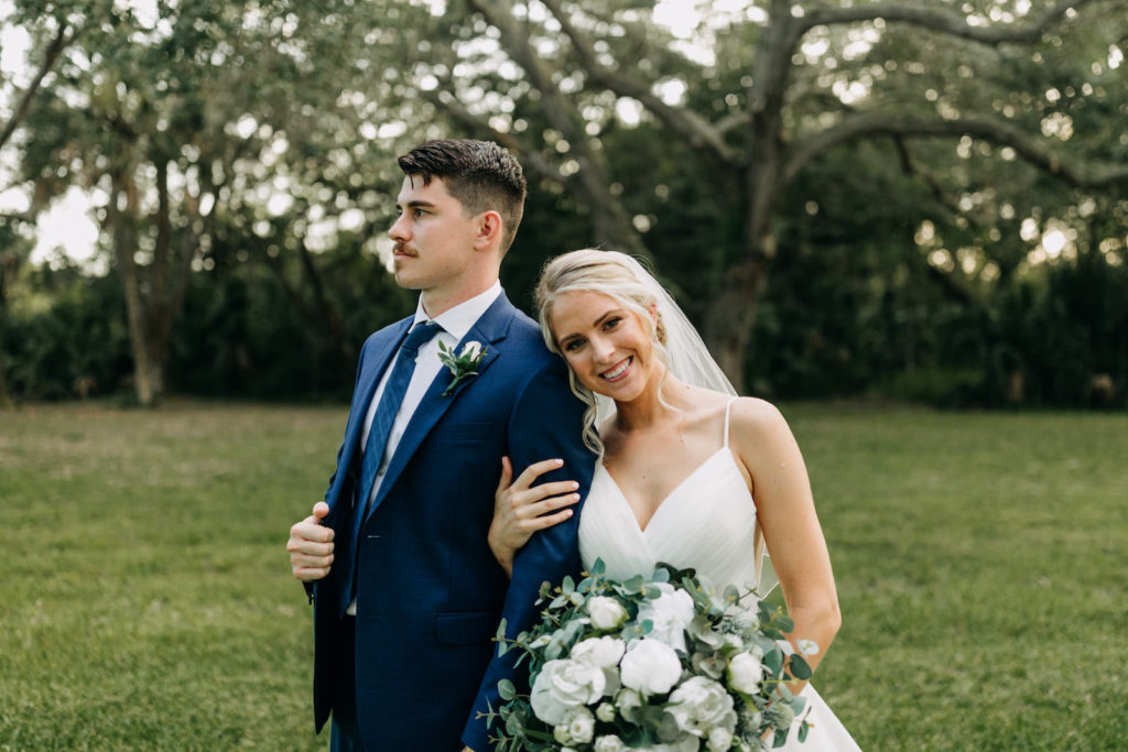 Classic Bride with Hair in Bun and Veil in V Neckline Wedding Dress Holding Eucalyptus Greenery and White Floral Bouquet with Groom in Blue Suit | Tampa Bay Wedding Photographer Amber McWhorter Photography | Wedding Hair and Makeup Femme Akoi Beauty Studio