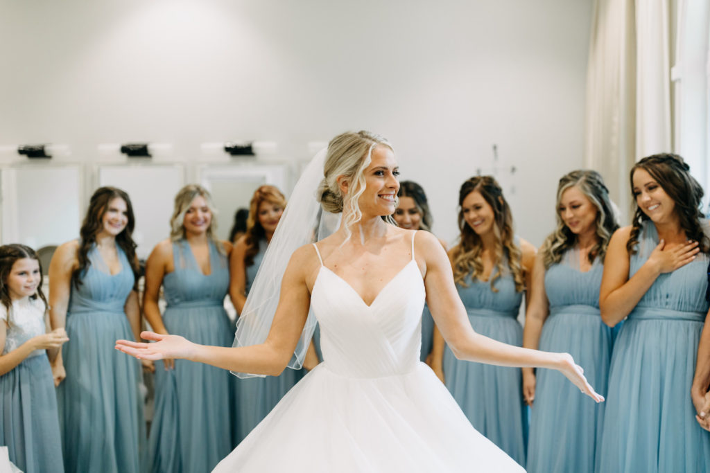 Classic Bride with Hair in Bun and Veil in Full Skirt V Neckline Wedding Dress First Look with Bridesmaids in Matching Dusty Blue Dresses | Tampa Bay Wedding Photographer Amber McWhorter Photography | Wedding Hair and Makeup Femme Akoi Beauty Studio