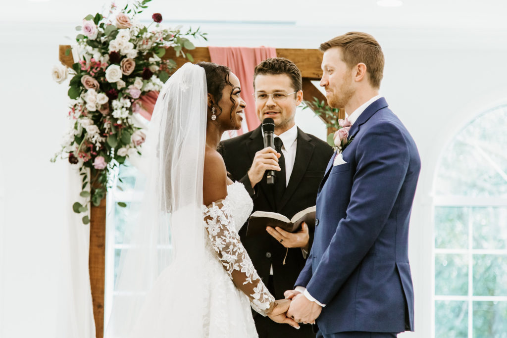 Bride and Groom Exchanging Vows during Indoor Church Ceremony at St. Pete Wedding Venue Harborside Chapel | Wood Arch with Draping and Natural Floral Arrangement Swag with Pink and White Roses and Eucalyptus Greenery | Lace and Tulle Ballgown Bridal Gown Wedding Dress with Illusion Lace Long Sleeves | Navy Groom Suit