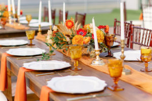 Modern Boho Inspired Florida Wedding Reception and Decor | Long Wooden Feasting Table with Orange Linen Table Runner with Long White Candles and Gold Candleholder, White Chargers, Acrylic Table Number, Yellow Drinking Goblets, Red Flowers, Marigolds Centerpiece | Tampa Bay Wedding Planner Coastal Coordinating