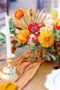 Modern Boho Inspired Florida Wedding Reception and Decor | Long Wooden Feasting Table with Orange Linen Table Runner with Long White Candles and Gold Candleholder, White Chargers, Acrylic Table Number, Yellow Drinking Goblets, Red Flowers, Marigolds Centerpiece | Tampa Bay Wedding Planner Coastal Coordinating