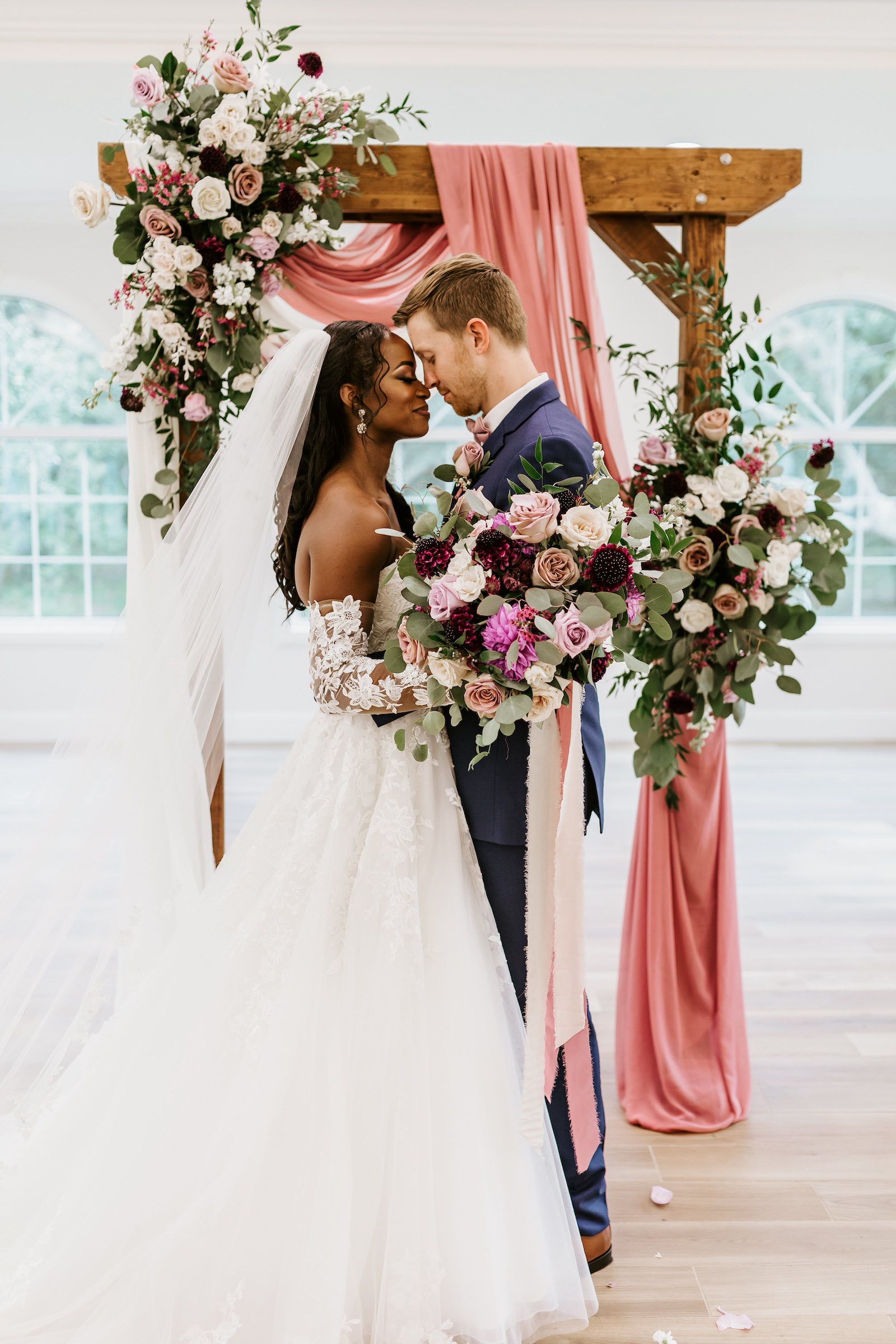 Bride and Groom Indoor Church Portrait at St. Pete Wedding Venue Harborside Chapel | Wood Arch with Draping and Natural Floral Arrangement Swag with Pink and White Roses and Eucalyptus Greenery | Lace and Tulle Ballgown Bridal Gown Wedding Dress with Illusion Lace Long Sleeves | Navy Groom Suit | Bridal Bouquet with Ivory Pink and Burgundy Maroon Roses with Eucalyptus Greenery and Cascading Pink Velvet Ribbons