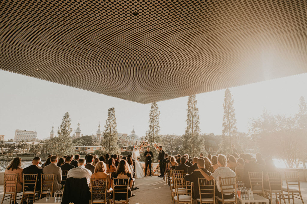 Outdoor Downtown Tampa Waterfront Wedding Ceremony with Gold Chiavari Chairs and Round Arch Floral Arrangement with Peach Salmon Coral Roses White Hydrangea and Eucalyptus Greenery
