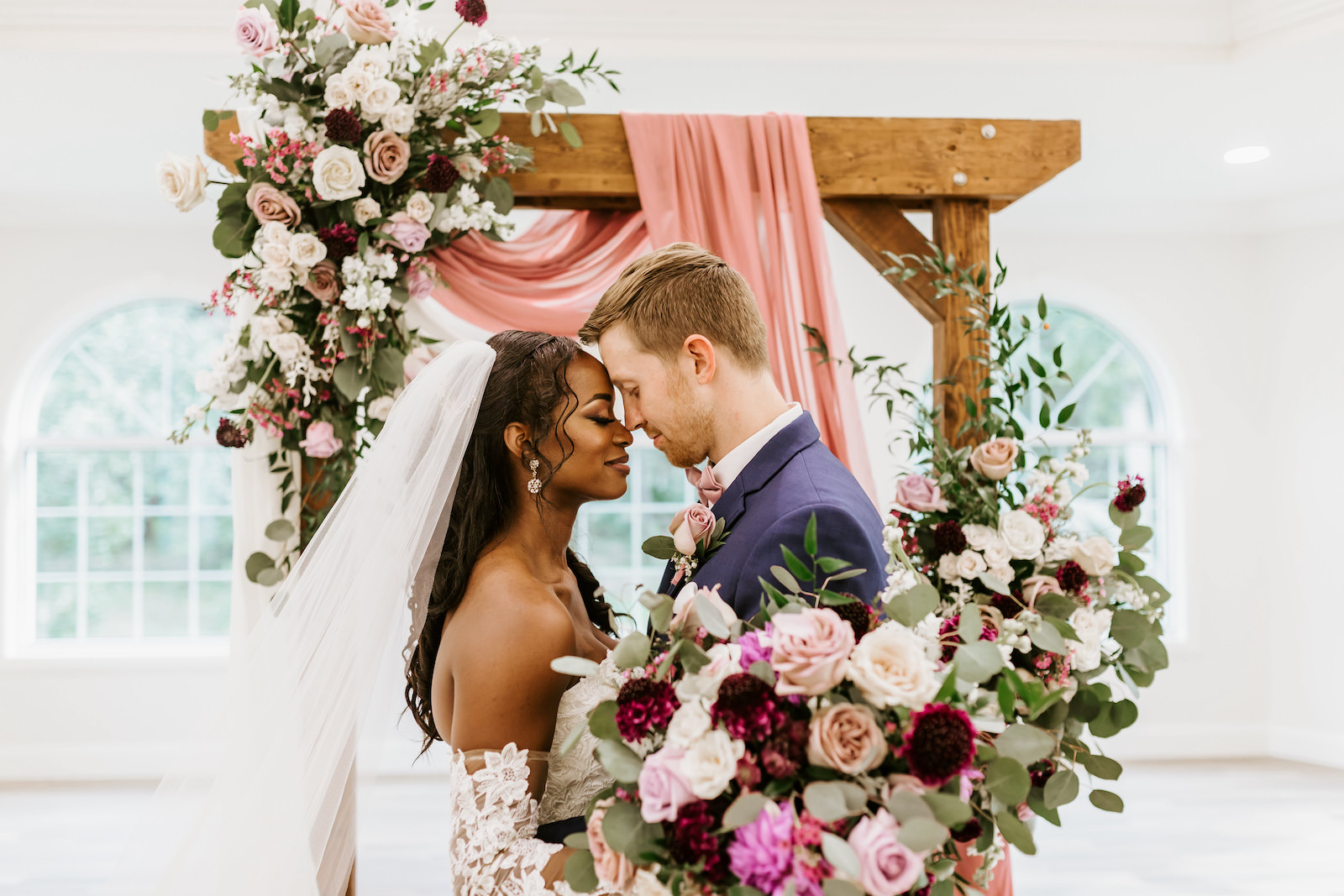 Bride and Groom Indoor Church Portrait at St. Pete Wedding Venue Harborside Chapel | Wood Arch with Draping and Natural Floral Arrangement Swag with Pink and White Roses and Eucalyptus Greenery | Lace and Tulle Ballgown Bridal Gown Wedding Dress with Illusion Lace Long Sleeves | Navy Groom Suit | Bridal Bouquet with Ivory Pink and Burgundy Maroon Roses with Eucalyptus Greenery and Cascading Pink Velvet Ribbons