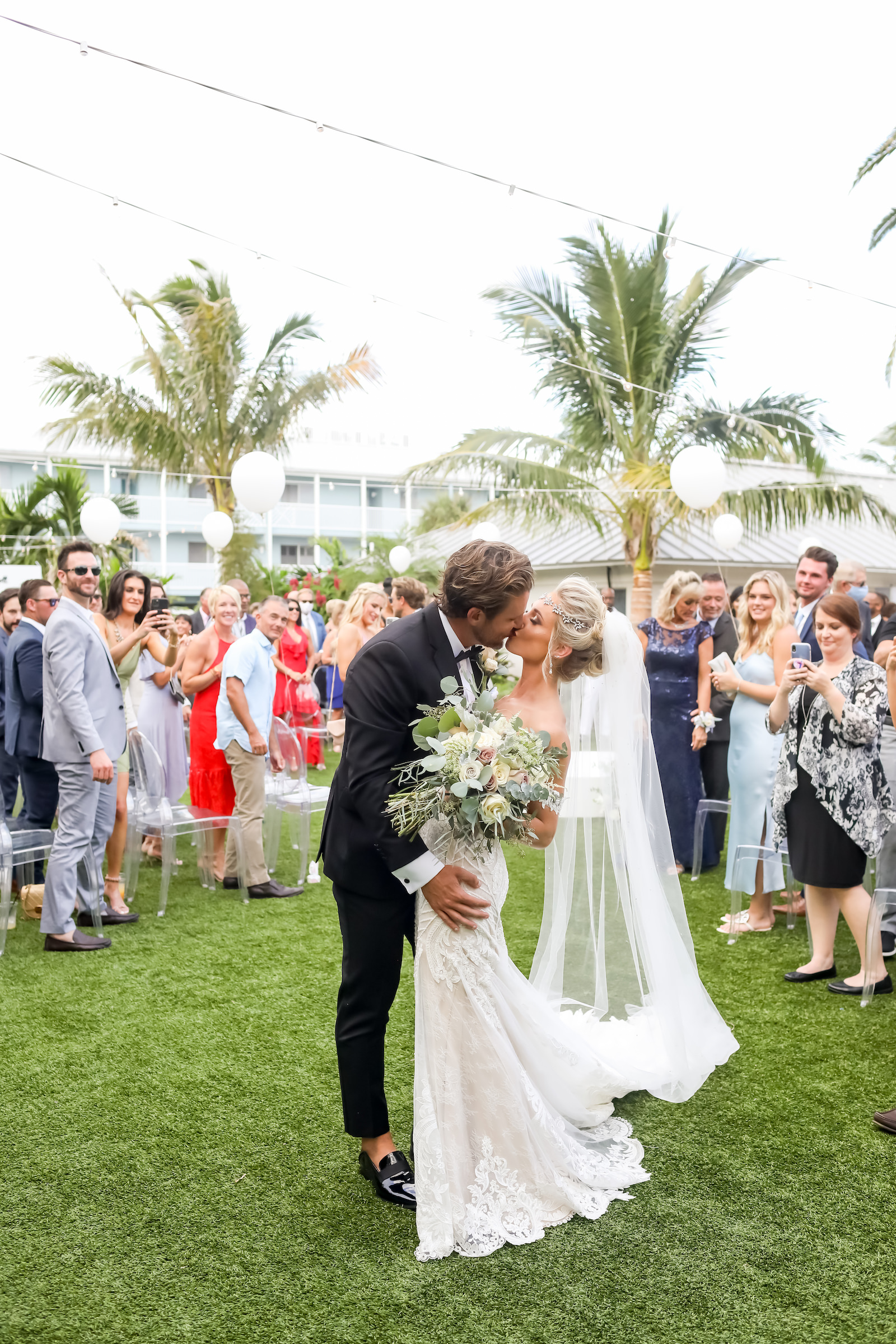 Florida Bride and Groom Kiss At End of the Aisle, Surrounded by Family and Friends on Acrylic Ghost Chairs, Tropical Elegant Inspired Floral Arrangements with Whtie and Ivory Flowers with Greenery | Sarasota Wedding Planner Kelly Kennedy Weddings | Florida Wedding Photographer Lifelong Photography Studio