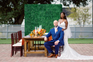 Downtown St. Petersburg Bride and Groom, Modern Boho Florida BrideWearing White Lace Mermaid Style Dress, Groom in Dark Blue Suit, Long Wooden Feasting Table with Vibrant Yellow and Red Floral Centerpiece, Tall White Candles and Gold Candlesticks, Wooden Chiavari Chairs, Greenery Wall Backdrop | Tampa Bay Wedding Planner Coastal Coordinating
