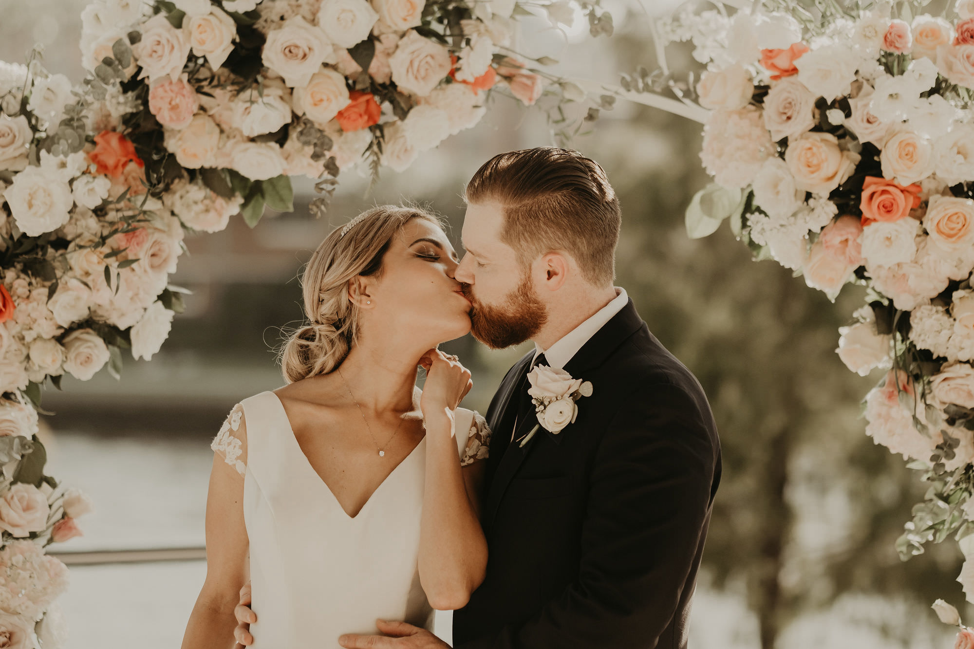 Outdoor Bride and Groom Portrait Kiss in front of Floral Arrangement with Peach Salmon Coral Roses White Hydrangea and Eucalyptus Greenery