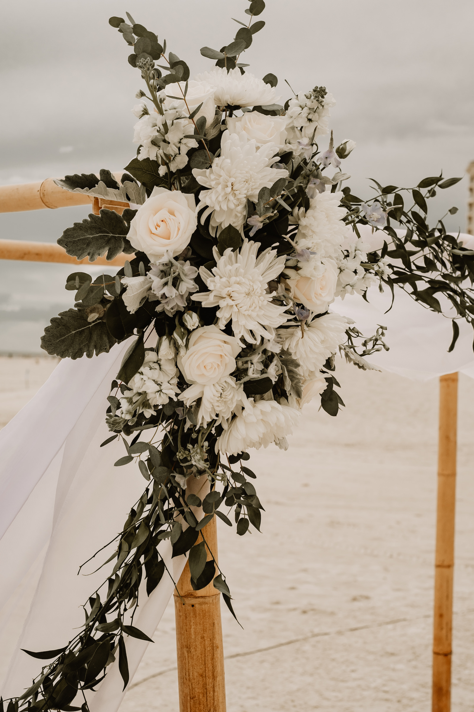 Beach Wedding Ceremony Bamboo Arch with Sheer Draping and Greenery Floral Arrangement Tie Backs with White Roses Chrysanthemums and Stock