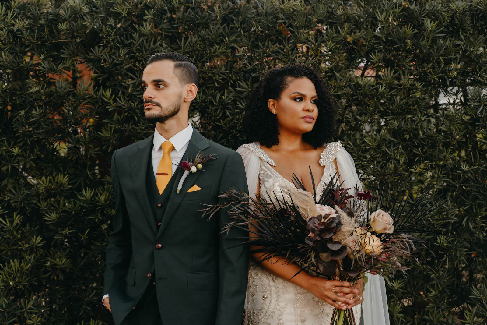 Outdoor Bride and Groom Portrait in Tampa Wedding Venue Hyde House | Illusion Lace Embroidered Beaded V Neck Wedding Dress Bridal Gown with Sheer Tulle Cape Sleeves by Designer Amalia Carrara Bridal | Groom in Classic Black Tux Suit with Gold Tie | Loose Wild Boho Bridal Bouquet with Deep Red Burgundy and Blush Roses