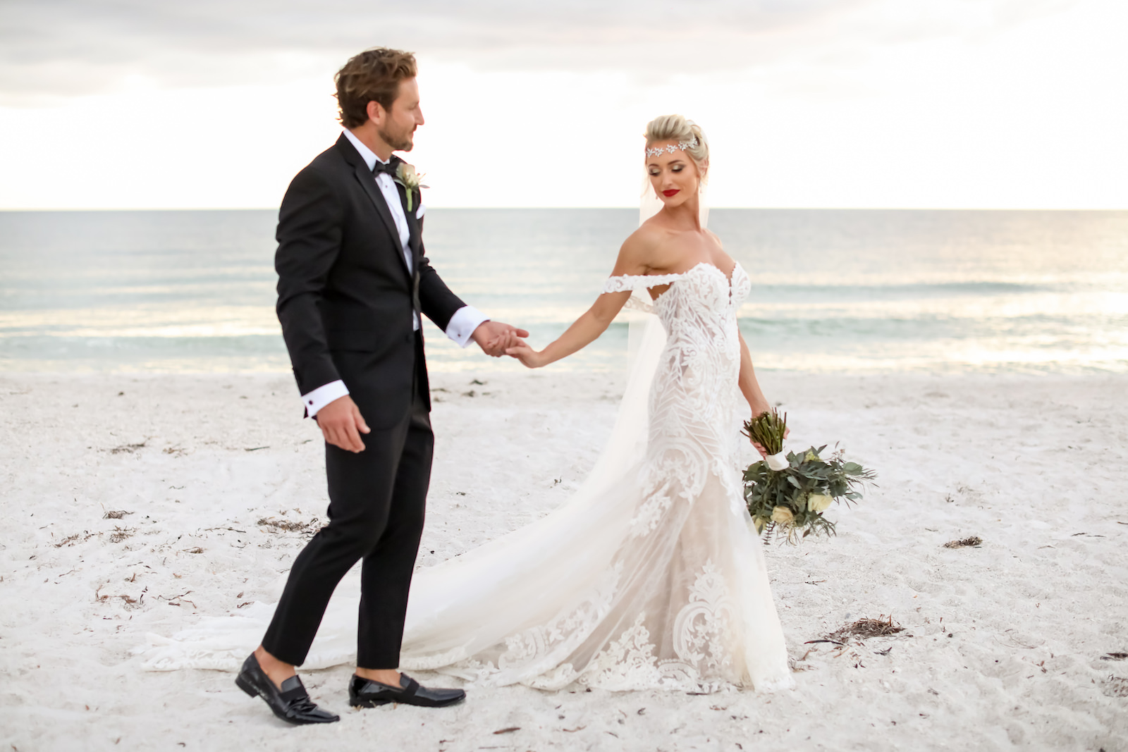 Elegant Sarasota Bride and Groom Walking on Anna Marie Island Beach at Sunset, Wearing Vintage Crystal Headpiece with White Fit and Flare Wedding Dress and Off the Shoulder Lace Sleeves, Holding Lush Ivory Floral Bouquet with Greenery, Groom in Classic Black Tuxedo | Florida Wedding Photographer Lifelong Photography Studio | Kelly Kennedy Weddings and Events